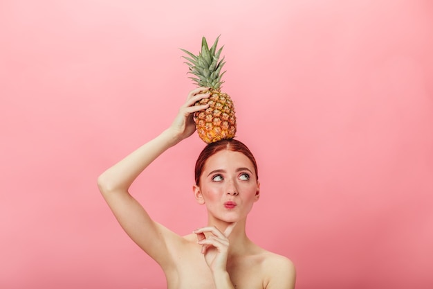 Mujer desnuda pensativa con piña. Chica caucásica de jengibre con frutas exóticas y mirando a otro lado.