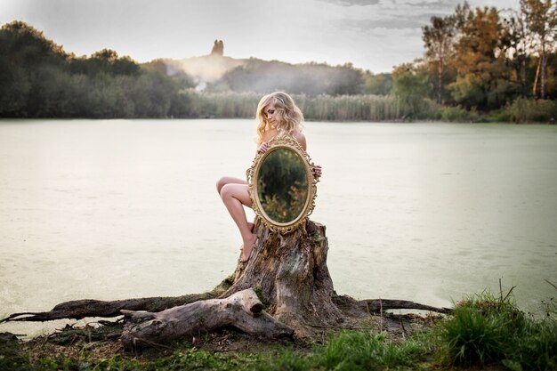 La mujer desnuda rubia sostiene un espejo que se sienta antes de un lago cubierto con la niebla