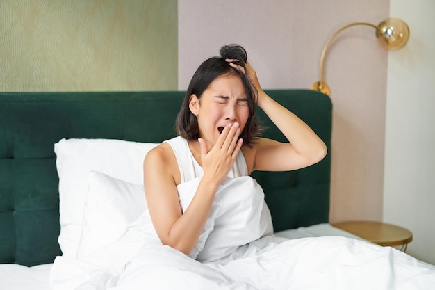 Mujer despertándose en la cama bostezando y haciendo muecas temprano en la mañana después de dormir sentada en el dormitorio