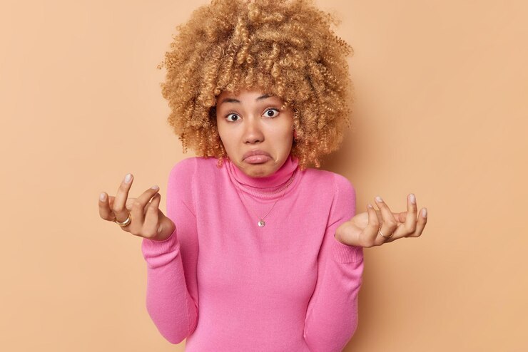 Foto gratuito la mujer despistada se siente perpleja, se encoge de hombros, los hombros, los bolsos, los labios, se ve vacilante, toma una decisión, usa un cuello de polo rosa casual aislado sobre fondo marrón. modelo femenino confundido con cabello rizado sin darse cuenta