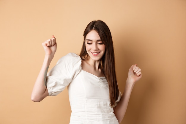 Mujer despreocupada bailando y divirtiéndose cerrar los ojos y sonriendo de pie sobre fondo beige