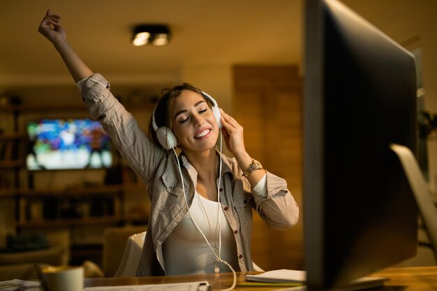 Mujer despreocupada con los ojos cerrados disfrutando de la música con los auriculares mientras usa la computadora por la noche en casa