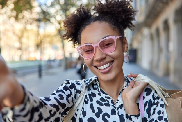 Una mujer despreocupada positiva con dos moños lleva gafas de sol rosas de moda y una blusa lleva poses de bolsas de la compra al aire libre durante el día. Compradora femenina sonriente posa para hacer selfie en ciudad