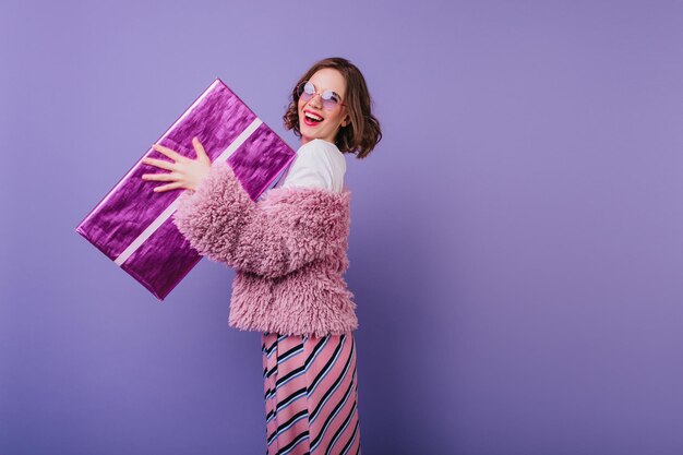 Mujer despreocupada que se ríe sosteniendo una caja de regalo brillante sobre fondo púrpura Foto interior de una deslumbrante cumpleañera con el pelo ondulado posando con un regalo