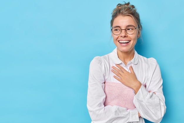 Una mujer devota feliz mantiene la mano en el pecho, se ve alegremente lejos, usa gafas redondas, camisa blanca formal, obtiene poses de noticias positivas contra el fondo azul con espacio en blanco para su promoción