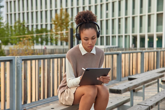 mujer dibuja bocetos en el panel táctil con lápiz óptico usa auriculares inalámbricos tiene habilidades creativas usa ropa ordenada posa al aire libre frente al entorno urbano