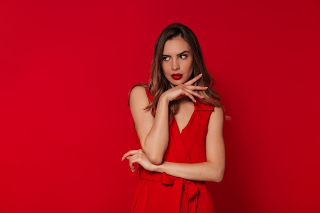 Mujer dichosa con maquillaje de noche con vestido rojo posando sobre pared roja