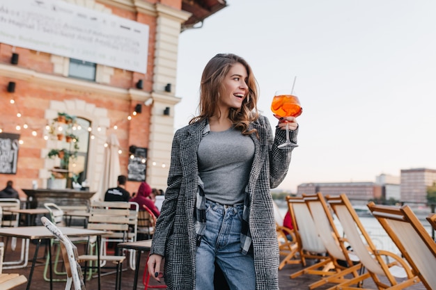 Mujer dichosa en traje casual levantando copa con cóctel de naranja en el fondo de la ciudad
