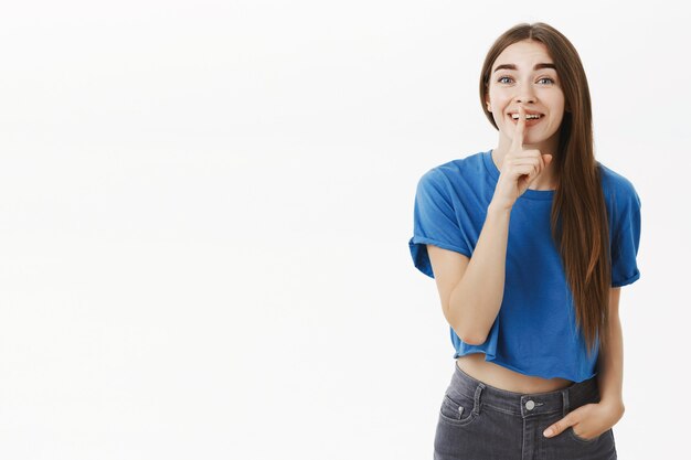 Mujer diciendo shh pidiendo silencio, chismeando haciendo gesto de silencio con el dedo índice sobre la boca, sonriendo con alegría sobre la pared gris