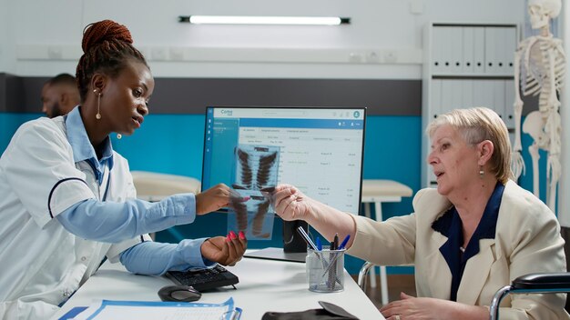 Mujer con discapacidad física analizando la exploración de rayos X con el médico en la visita de control en el gabinete médico. Viejo usuario de silla de ruedas que mira los resultados del diagnóstico de radiografía de huesos, atención médica.