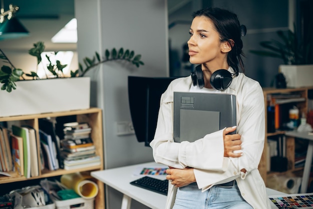 Mujer diseñadora con auriculares musicales de pie en la oficina con carpeta