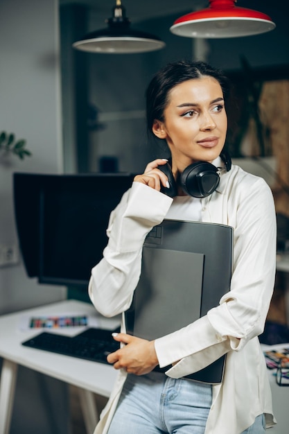 Mujer diseñadora con auriculares musicales de pie en la oficina con carpeta