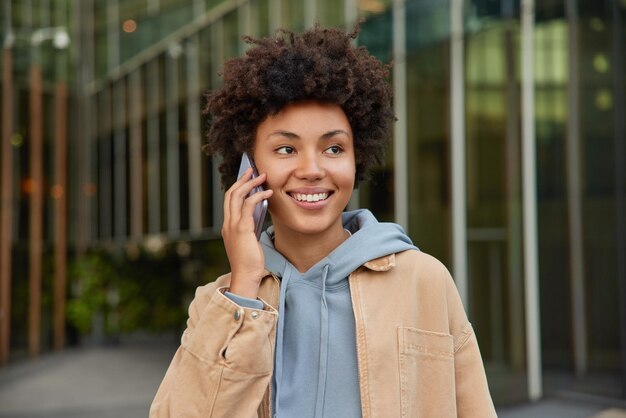 La mujer disfruta de la comunicación celular sonríe positivamente vestida casualmente plantea al aire libre hace una llamada telefónica