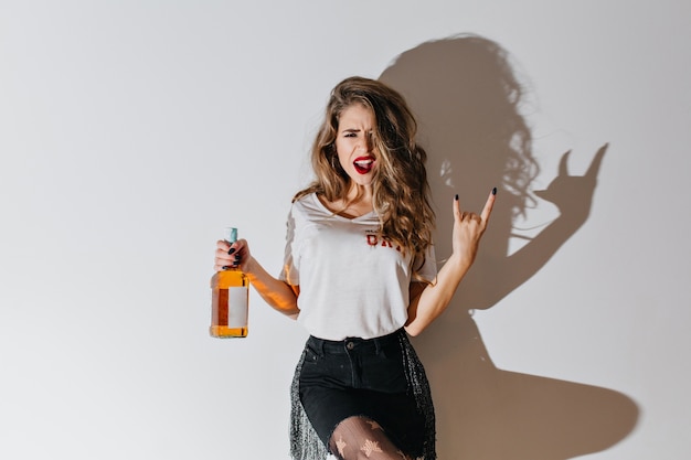 Mujer disgustada en pantimedias posando con botella de brandy en la pared blanca