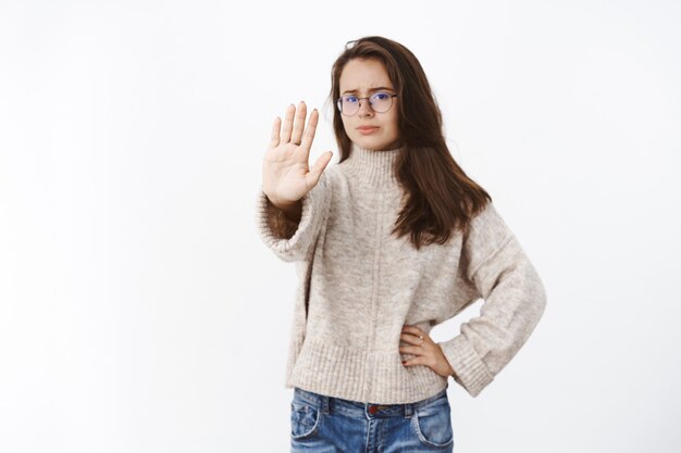 Mujer disgustada que expresa renuencia y disgusto extendiendo el brazo en gesto de parada frunciendo el ceño por disgusto y juicio rechazando y prohibiendo algo dudoso sobre la pared gris.