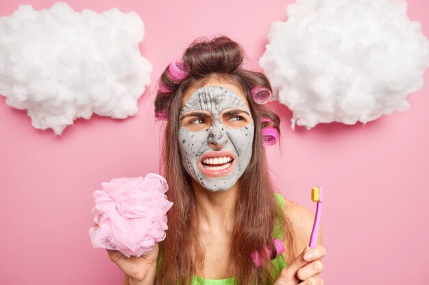 Mujer disgustada sonríe con satisfacción muestra los dientes sostiene el cepillo de dientes va a tomar una ducha aplica mascarilla de arcilla para refrescar la piel hace peinado con rodillos se somete a procedimientos de belleza quiere lucir muy hermosa