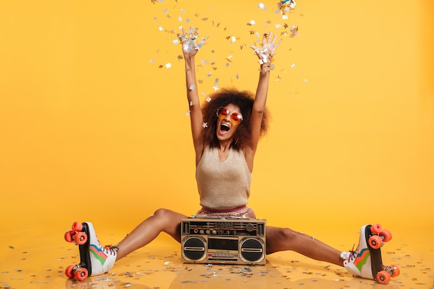 Mujer disko africana emocional en ropa retro y patines tirando confeti mientras está sentado con boombox