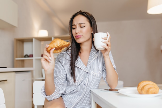 Mujer divertida con pelo negro lacio comiendo croissant durante el desayuno en acogedores apartamentos