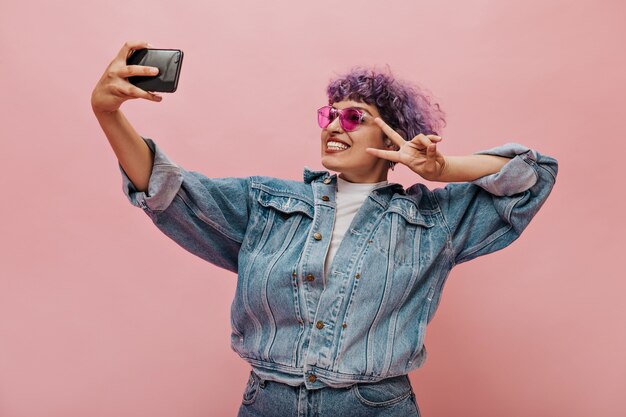 Mujer divertida con el pelo rizado en gafas toma una foto y muestra el signo de la paz. Señora brillante en chaqueta de gran tamaño posando.