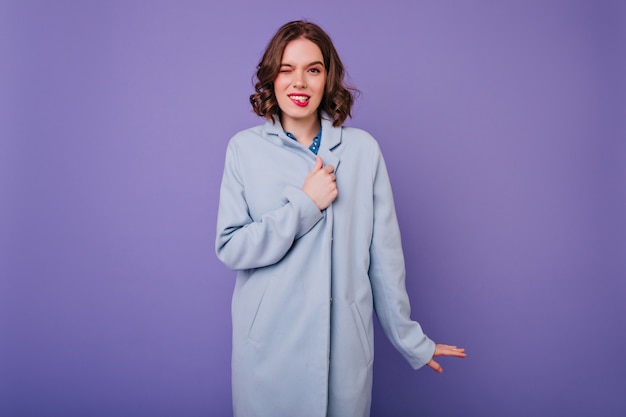 Mujer divertida con pie de pelo ondulado en abrigo azul. Maravillosa chica caucásica con maquillaje brillante haciendo muecas en la pared púrpura.