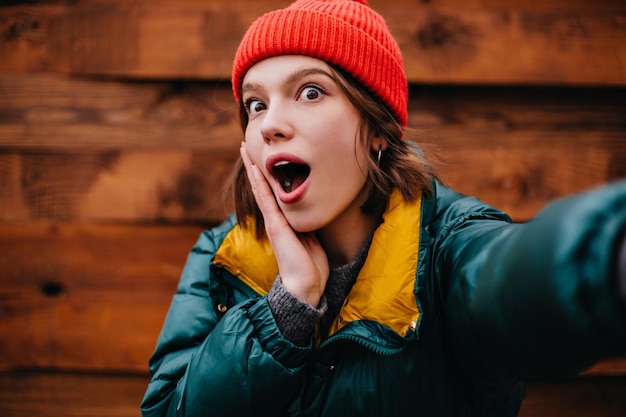 Mujer divertida con ropa de invierno elegante mira sorprendida a la cámara sobre fondo de madera Chica de pelo corto con sombrero rojo hace selfies