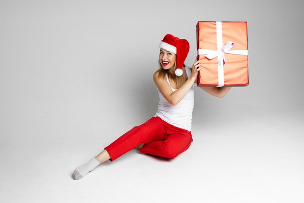 Mujer divertida con sombrero de Navidad se sienta con una gran caja roja y trata de escuchar lo que hay dentro, imagen aislada sobre fondo blanco.