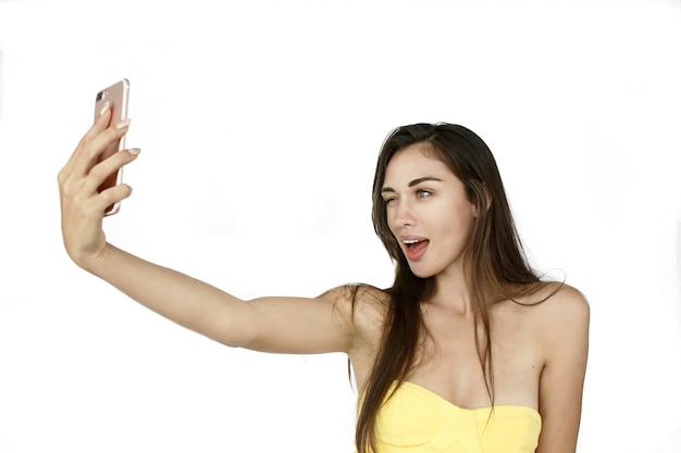 La mujer divertida toma el selfie en su teléfono que se coloca en el fondo blanco