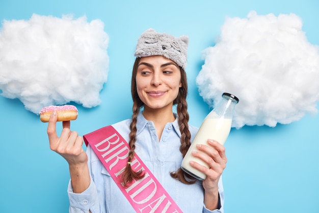 Mujer con dos coletas mira sabroso donut glaseado que va a comer con leche fresca le gustan los deliciosos postres dulces viste ropa doméstica aislado en azul