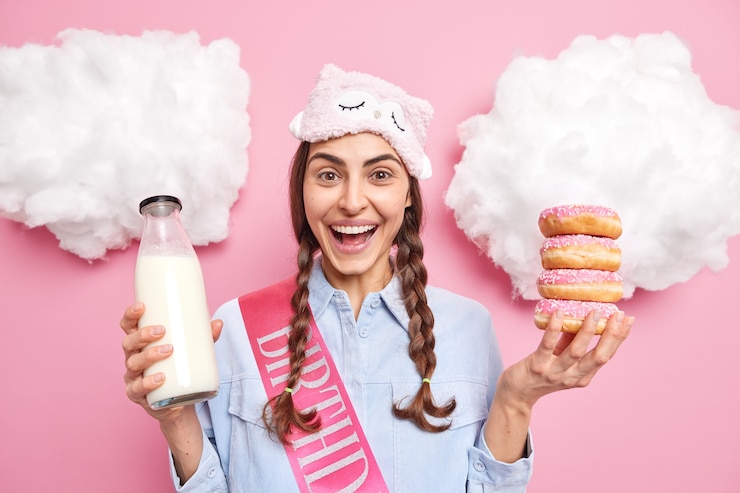 Foto gratuito mujer con dos coletas sonríe ampliamente sostiene deliciosas donas y leche tiene un estado de ánimo feliz, usa una venda en la frente aislado en rosa