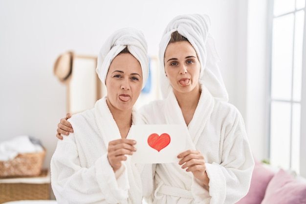 Mujer e hija de mediana edad con bata de baño sosteniendo una tarjeta de corazón sacando la lengua feliz con una expresión graciosa.