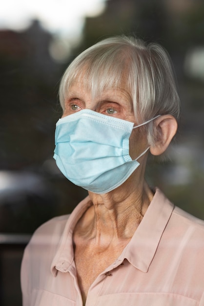 Mujer de edad avanzada con máscara médica mirando a través de la ventana de cristal