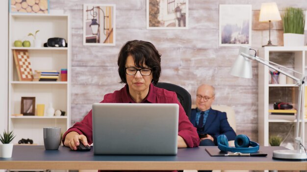 Mujer de edad avanzada tomando un sorbo de café mientras navega en la computadora portátil. Anciano relajante en el sofá en el fondo.