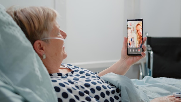 Mujer de edad sosteniendo teléfono inteligente con videollamada para comunicación remota con sobrina e hija. Paciente enfermo que usa una conferencia en línea para hablar con familiares mientras está acostado en la cama de la sala de hospital.