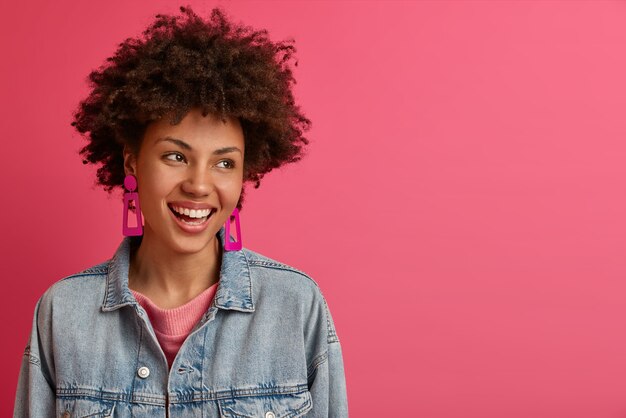 La mujer elegante y afortunada con peinado afro mira a un lado, sonríe positivamente, disfruta de la vida, se divierte con alguien, usa poses de chaqueta de mezclilla en el interior contra la pared rosada, espacio en blanco para su anuncio