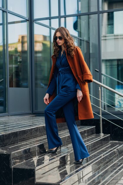 Mujer elegante y atractiva con caminar en la calle de negocios de la ciudad urbana vestida con abrigo marrón cálido y traje azul, estilo callejero de moda primavera otoño, con gafas de sol