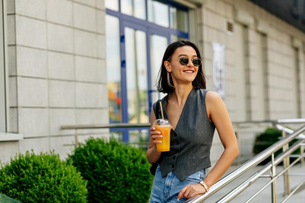 Una mujer elegante y atractiva que se ríe con un peinado corto lleva una chaqueta gris y jeans bebe limonada al aire libre en un día cálido y soleado