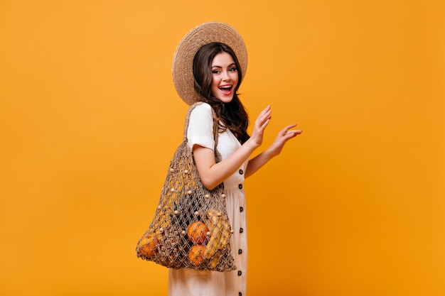 Mujer elegante con cabello ondulado posa con bolsa ecológica con frutas. Chica con sombrero de paja sonríe sobre fondo naranja.