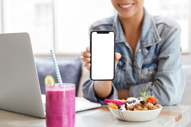 Mujer con elegante chaqueta vaquera en cafetería con pantalla de smartphone en blanco