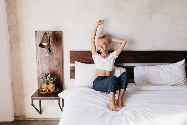 Mujer elegante complacida sentada en la cama. Señora caucásica pasar el fin de semana en su acogedor piso.