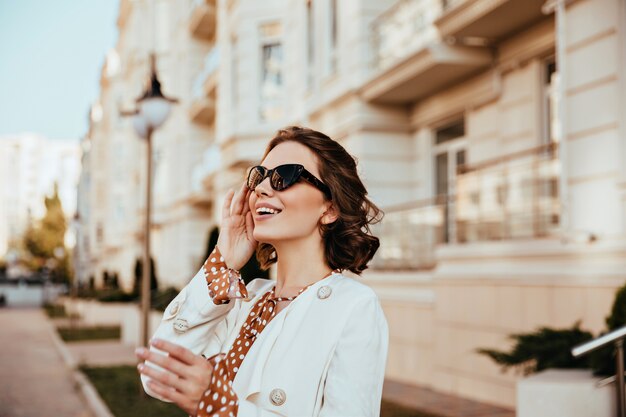 Mujer elegante dichosa posando cerca de gran edificio antiguo. Chica caucásica refinada de pie en el bakground de la ciudad borrosa en día de otoño