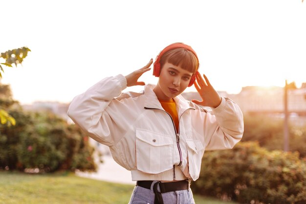 Mujer elegante escucha música en auriculares rojos al aire libre Hermosa chica de pelo corto en chaqueta caminando en el parque