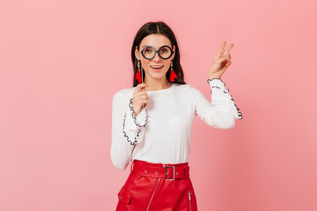 Mujer elegante en falda de cuero roja y pendientes brillantes muestra el signo de la paz sobre fondo rosa. Retrato de niña en anteojos redondos.