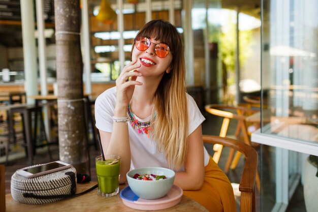 Mujer elegante feliz que come la comida sana que se sienta en el interior hermoso con las flores verdes