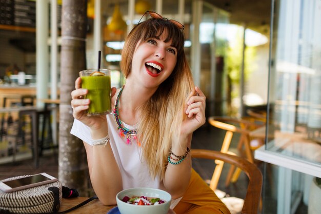 Mujer elegante feliz que come la comida sana que se sienta en el interior hermoso con las flores verdes