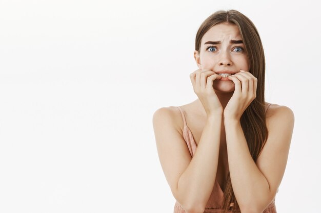 Mujer elegante femenina tímida e insegura asustada con cabello castaño mordiendo los dedos mirando asustado y asustado presa del pánico reaccionando exageradamente haciendo expresión de terror sobre la pared gris