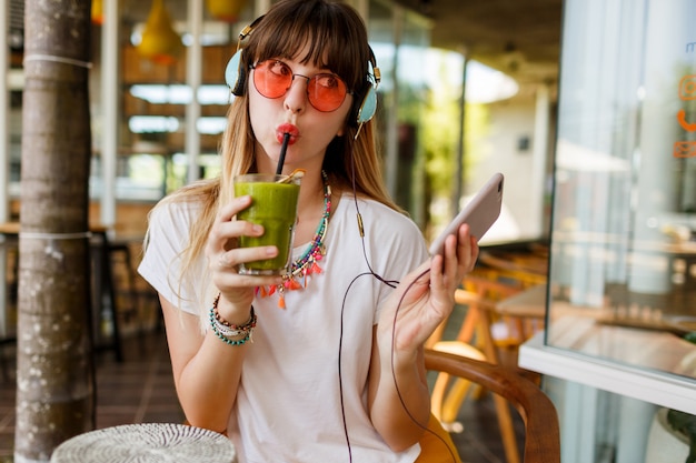 Mujer elegante en gafas de color rosa disfrutando de batido verde saludable, escuchando música por auriculares, sosteniendo el teléfono móvil.