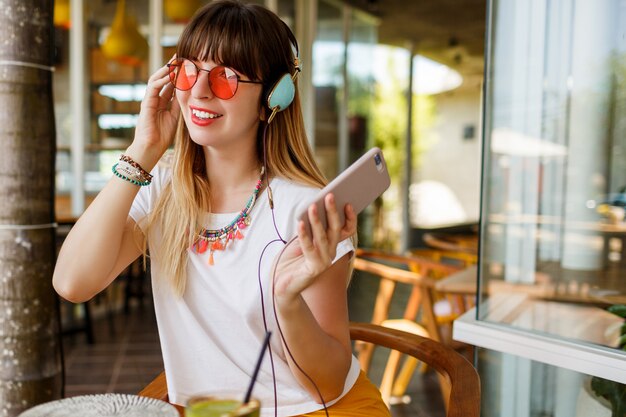 Mujer elegante en gafas de color rosa disfrutando de batido verde saludable, escuchando música por auriculares, sosteniendo el teléfono móvil.