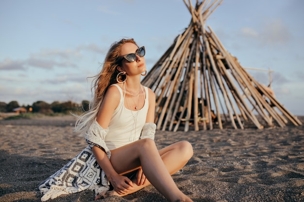 Mujer elegante en gafas de sol negras sentada en la playa y mirando al cielo.