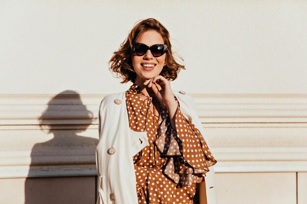 Mujer elegante inspirada en gafas de sol de pie junto a la pared. Tiro al aire libre de risa alegre niña con cabello castaño.