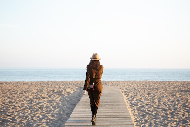 Mujer elegante irreconocible con sombrero, abrigo largo y bandolera caminando por el paseo marítimo